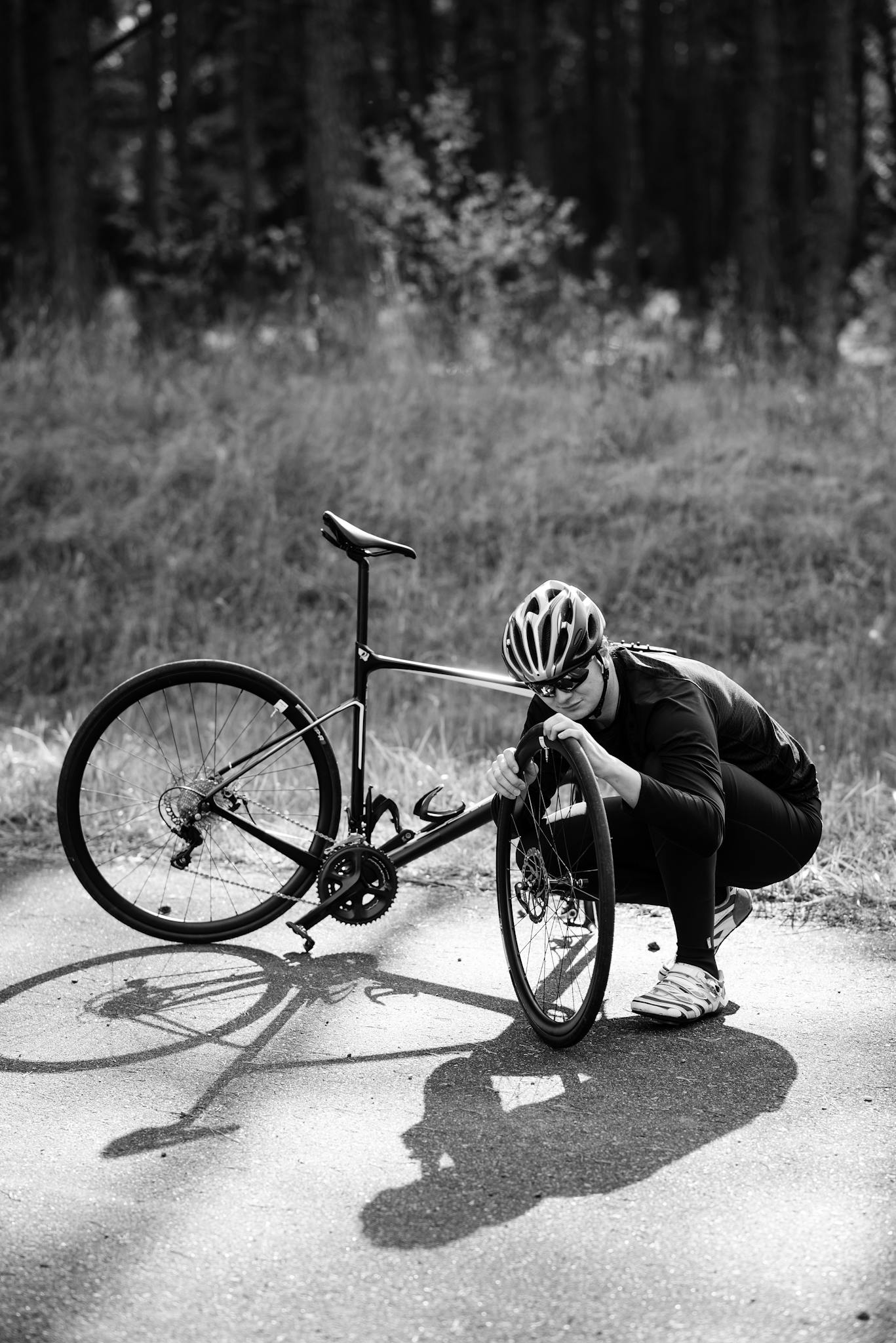 A cyclist in gear repairs their bike on a sunlit outdoor path, captured in black and white.