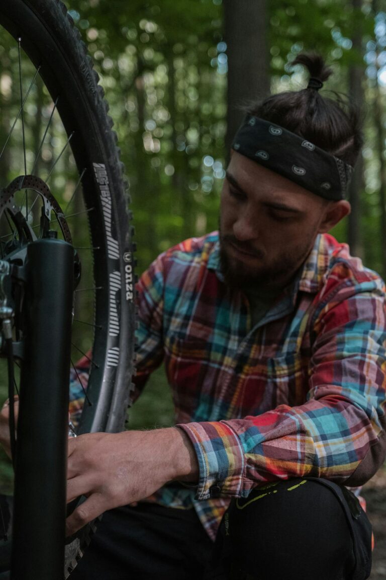 A man in a plaid shirt repairs a bicycle wheel in a forest.